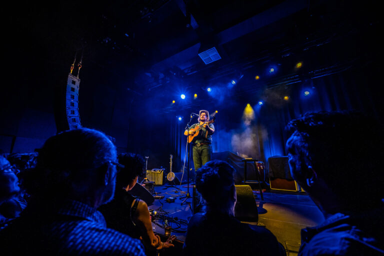 Een muzikant staat op het podium, omringd door blauwachtig licht, terwijl het publiek naar hem kijkt.