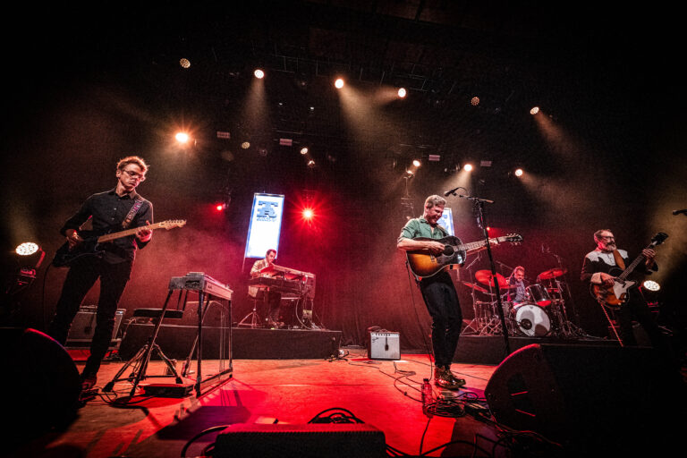Een band speelt live op een podium met kleurrijke verlichting en een energieke sfeer.