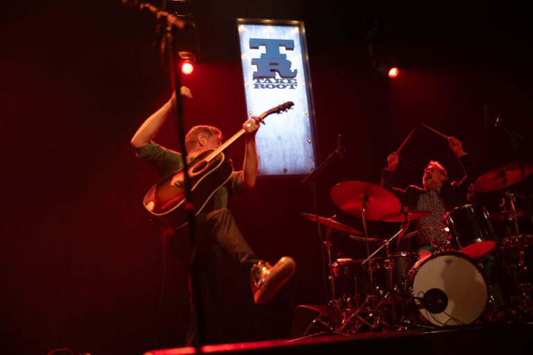 Een muzikant springt energiek met een gitaar op het podium, terwijl een drummer achter hem speelt.