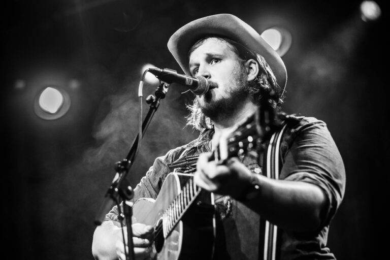 Een zanger met een gitaar, gekleed in een hoed, zingt in een podiumsetting met sfeerverlichting.