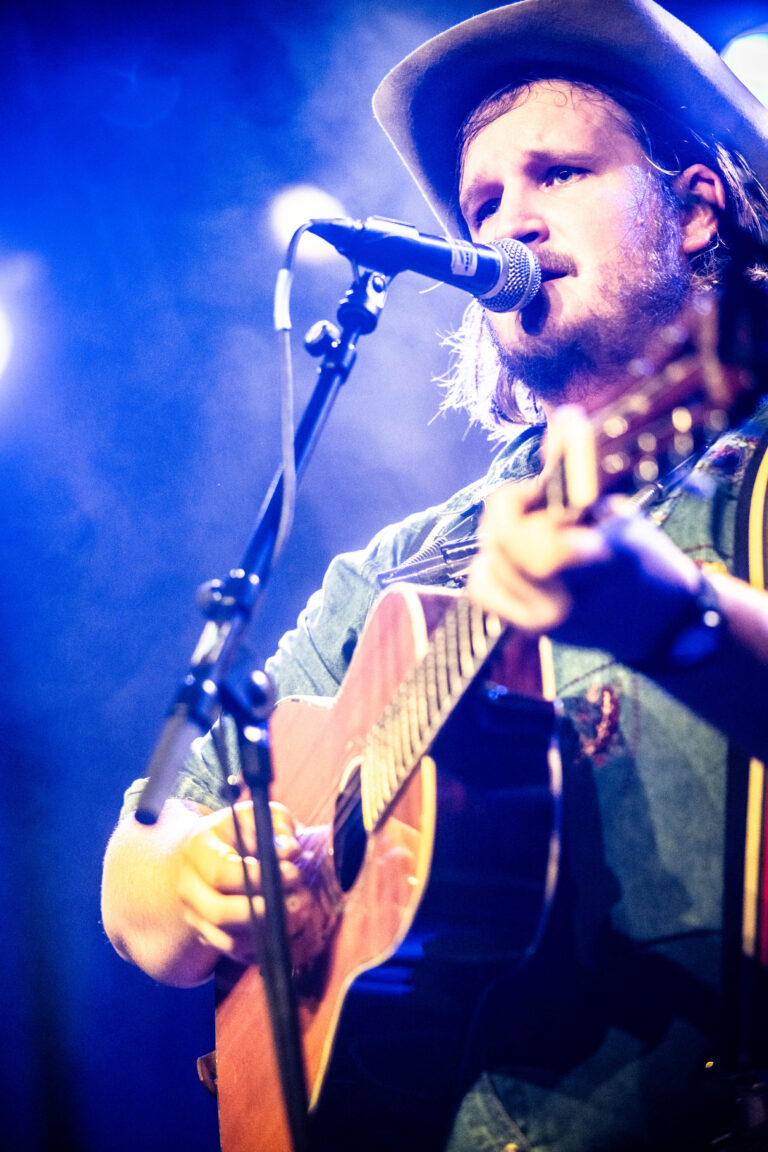 Een jonge man speelt gitaar en zingt in een microfoon, omringd door sfeervolle verlichting.