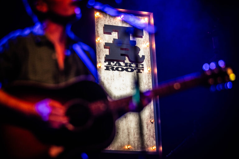 Een muzikant met een gitaar staat in de schijnwerpers, met op de achtergrond een verlichte spandoek met het logo van de muziekvenue.