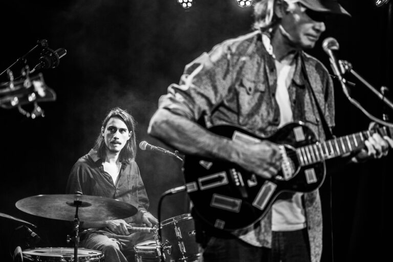 Een muzikant speelt gitaar terwijl een drummer op de achtergrond aandachtig kijkt.