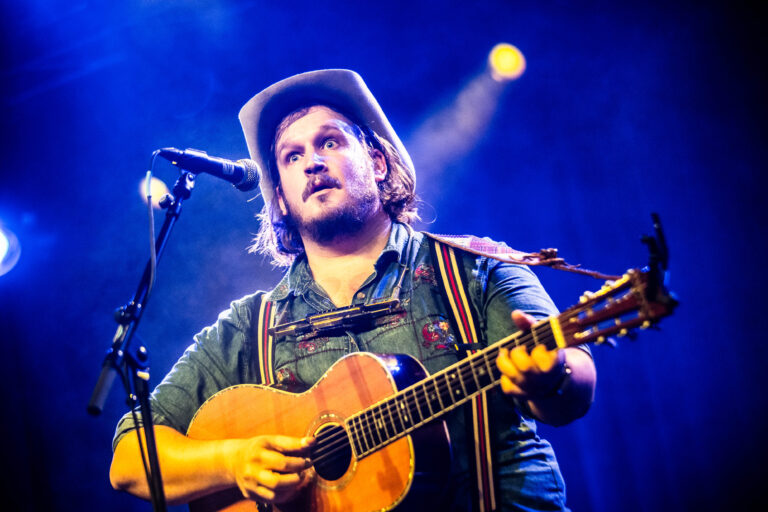 Een man met een hoed speelt gitaar op een podium, verlicht door kleurrijke lichten.