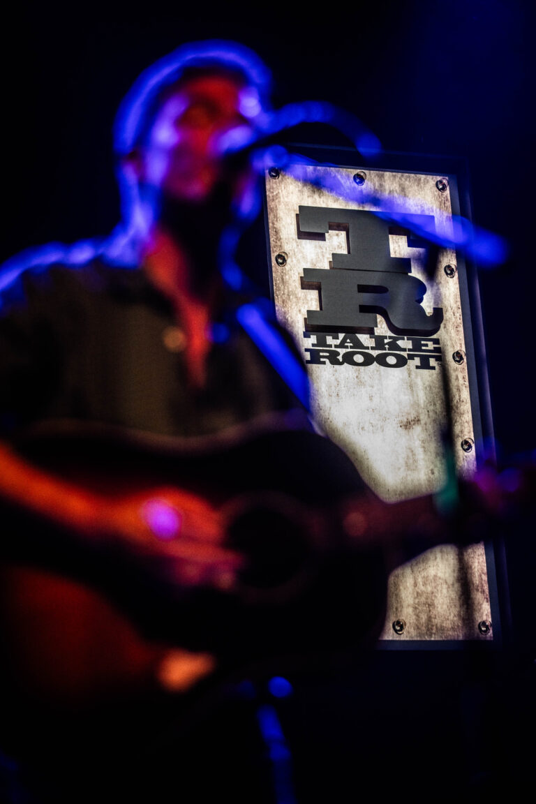 Een muzikant speelt gitaar voor een verlicht podium met een logo op de achtergrond.