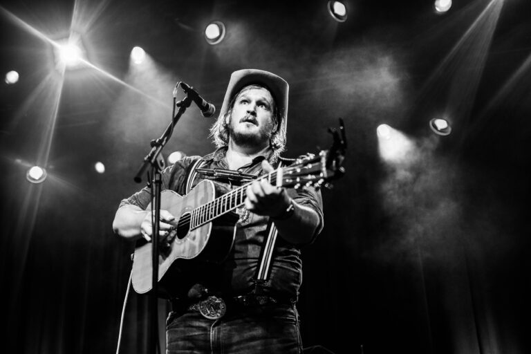 Een muzikant met een hoed speelt gitaar op een podium, omringd door lichteffecten.