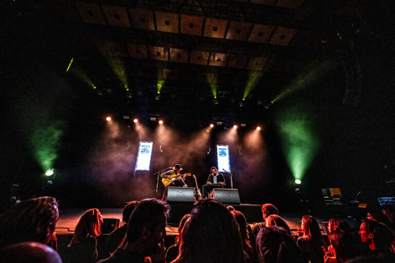 Twee muzikanten optreden op een podium met kleurrijke verlichting, voor een menigte.