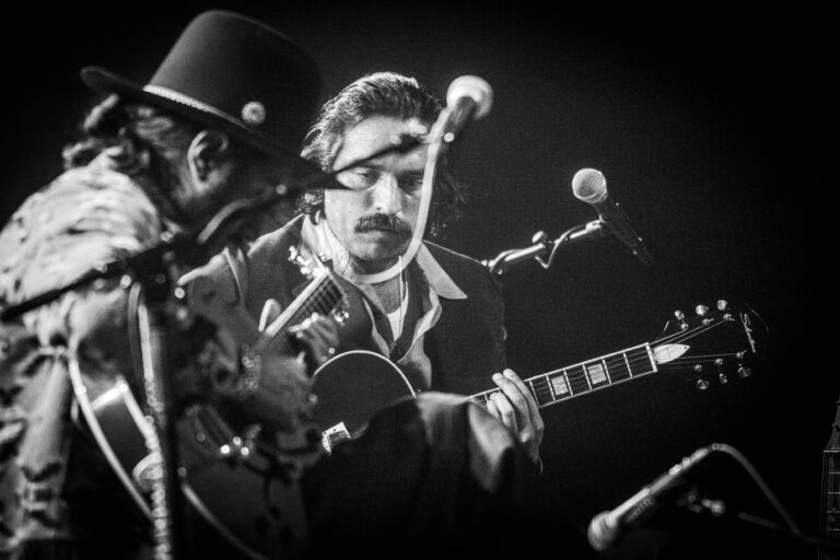 Een muzikant speelt gitaar op een podium, terwijl een andere persoon in de achtergrond zichtbaar is.