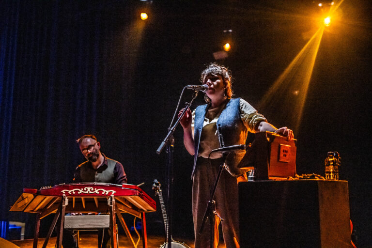 Een muzikant staat op het podium met een microfoon, terwijl een andere muzikant achter een keyboard zit, omringd door podiumverlichting.