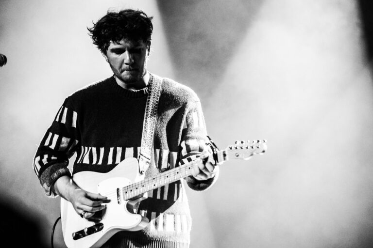 Een muzikant speelt op een witte gitaar, gekleed in een gestreept trui, met een dramatische achtergrondverlichting.