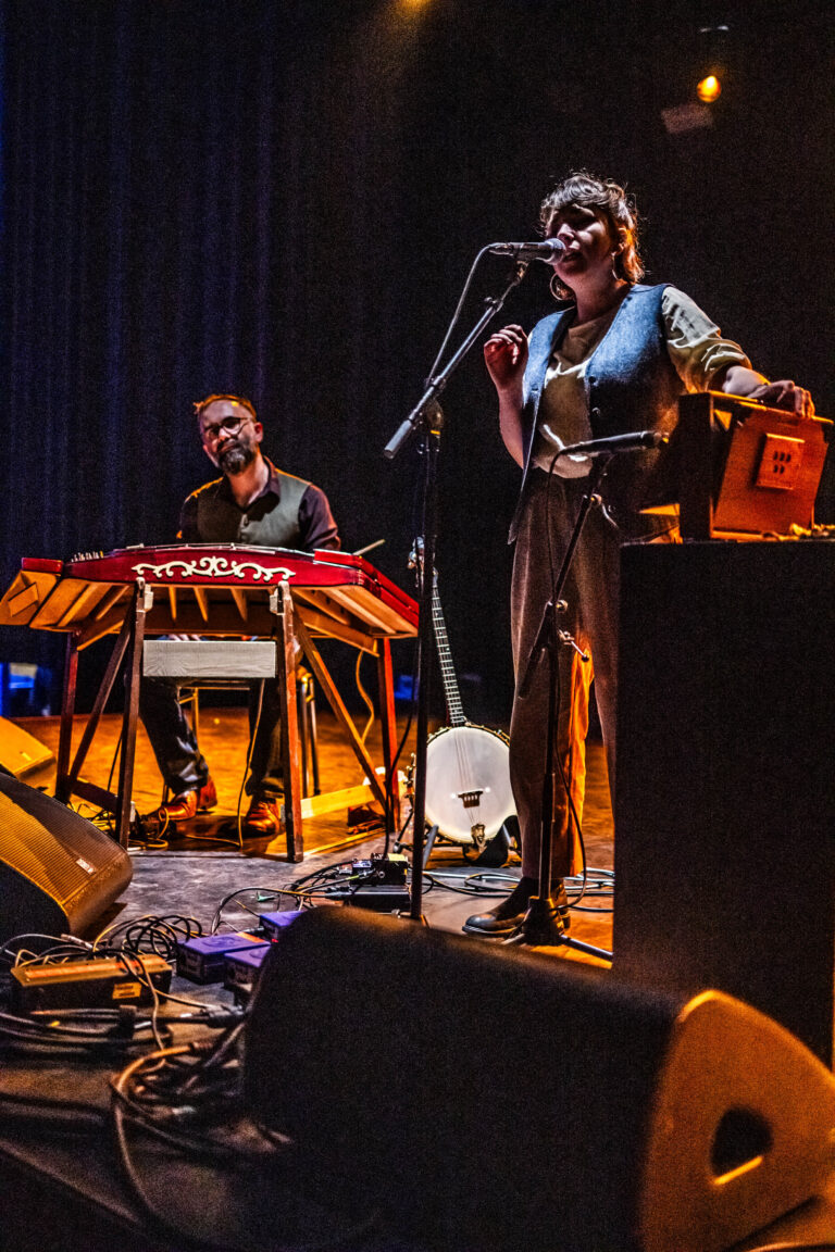 Een muzikant staat aan een microfoon terwijl een andere persoon op een keyboard speelt tijdens een liveoptreden.