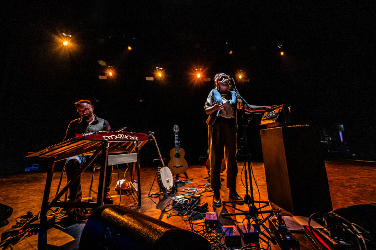 Een muzikant staat op het podium met een microfoon, terwijl een andere muzikant aan een keyboard speelt, omgeven door instrumenten en apparatuur.
