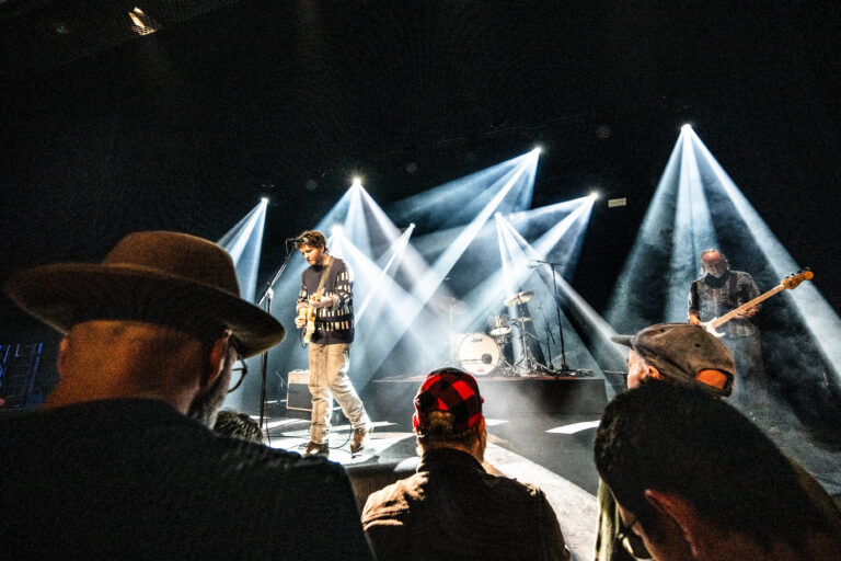 Een muzikant performt op een podium met heldere lichtstralen, terwijl een publiek van achteren toekijkt.