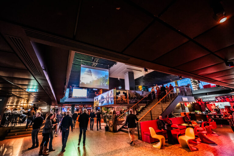 Een drukke zaal met kleurrijke verlichting, mensen en een trap die naar een bovenverdieping leidt, met een scherm aan de wand.