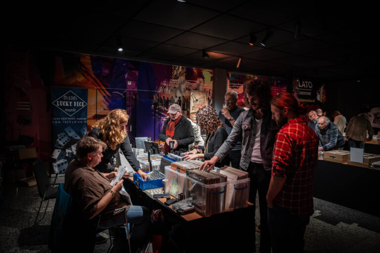 Een groep mensen bij een verkoopstand, druk in gesprek en bezig met het bekijken van producten in een donkere ruimte met kleurrijke achtergronden.