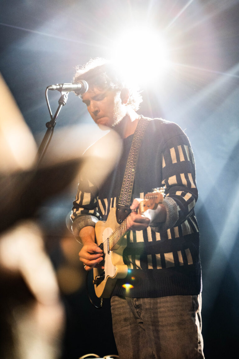 Een muzikant speelt gitaar op een podium, verlicht door fel lichtstralen.