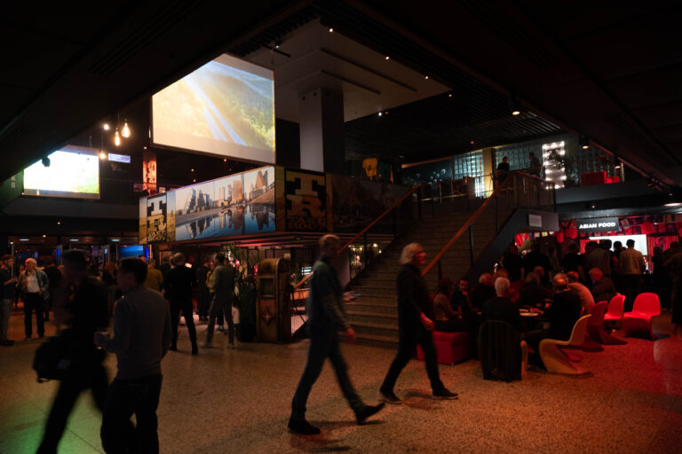Een drukke lobby met mensen die zich verplaatsen, een trappenhuis en een groot scherm bovenaan.