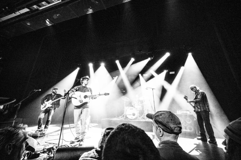 Een zwart-witfoto van een live optreden met muzikanten op het podium en sfeerverlichting.