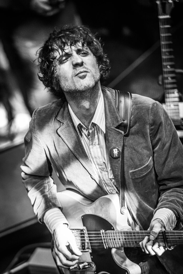 Een muzikant speelt gitaar in zwart-wit, met een expressieve uitstraling en een casual outfit.