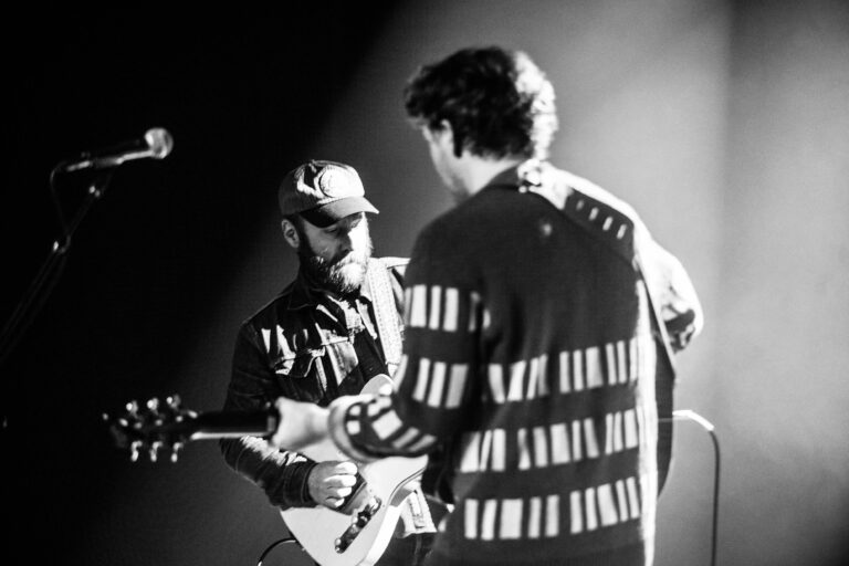 Twee muzikanten spelen samen op het podium, met een focus op de gitarist rechts in een gestreept truitje.