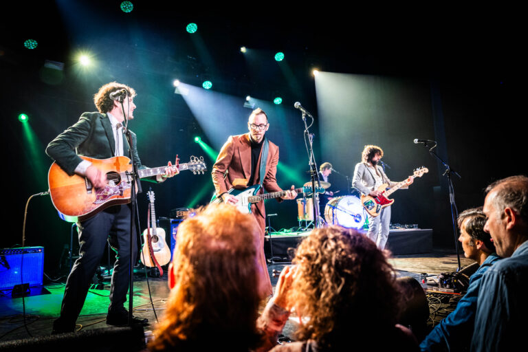 Een band speelt live op het podium met verschillende instrumenten onder kleurrijke verlichting.