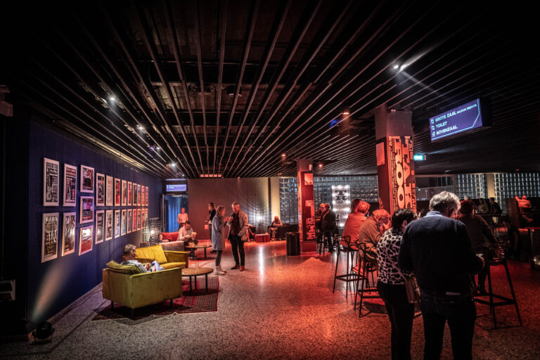 Een stijlvolle, sfeervolle ruimte met mensen die in gesprek zijn, kleurrijke kunst aan de muren en comfortabele zitplaatsen.