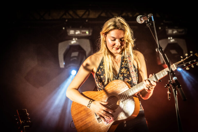 Een muzikante speelt gitaar op het podium, omringd door sfeerlicht.