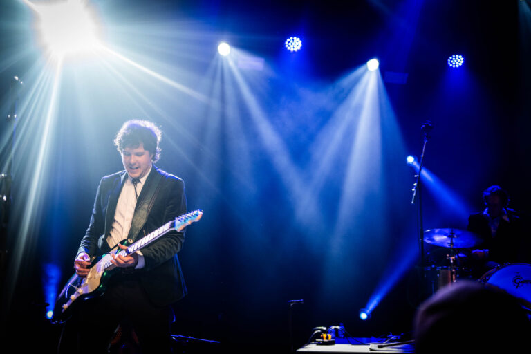 Een muzikant speelt gitaar op een podium, omringd door blauwe en witte lichten.