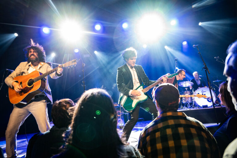 Een band speelt live op een podium met fel licht, terwijl het publiek geniet van de muziek.