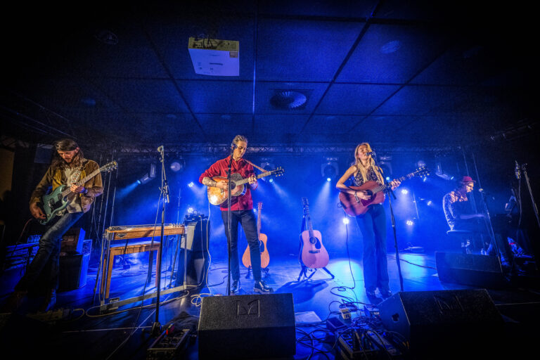 Een muzikale liveoptreden met vier artiesten die op verschillende instrumenten spelen, omringd door sfeervolle blauwe belichting.