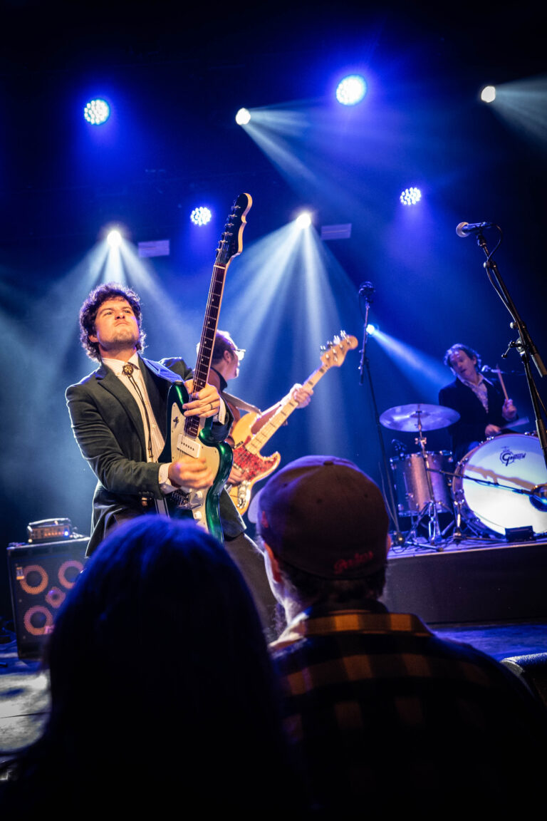 Een muzikant speelt gitaar op een podium, terwijl andere bandleden achter hem optreden onder kleurrijke lichteffecten.