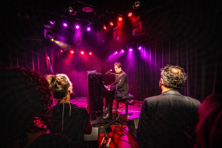 Een muzikant speelt piano op een podium met kleurrijke verlichting, terwijl enkele toeschouwers toekijken.