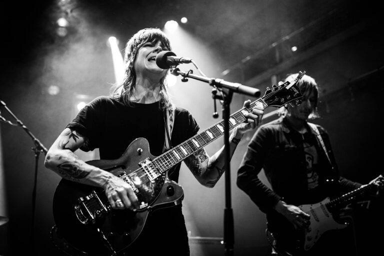 Een muzikant speelt gitaar en zingt op een podium, terwijl een andere muzikant op de achtergrond te zien is.