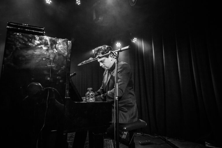 Een muzikant speelt piano op een podium, omringd door microfoons en een donkere achtergrond.