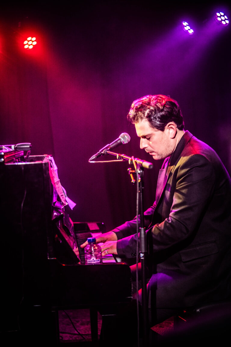 Een man in een zwart jasje speelt piano op een podium met kleureffecten van paars en rood.