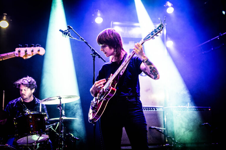 Een muzikant speelt gitaar op een podium met gekleurde lichten en een drumstel op de achtergrond.
