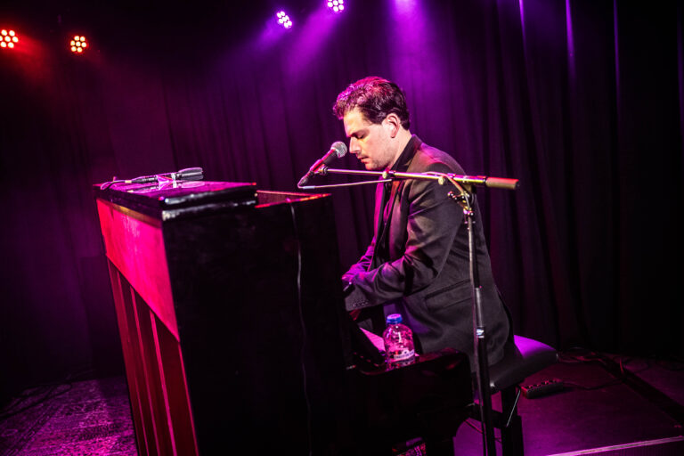 Een muzikant speelt piano op een podium, omgeven door paarse verlichting.