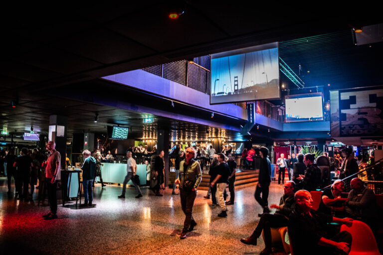 Een drukke bar of lounge met mensen die socialiseren en een moderne inrichting, inclusief schermen in de achtergrond.