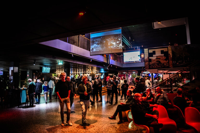 Een drukke evenementenruimte met mensen die zich verspreid over de vloer bevinden, verlicht door kleurrijke lichten en grote schermen.