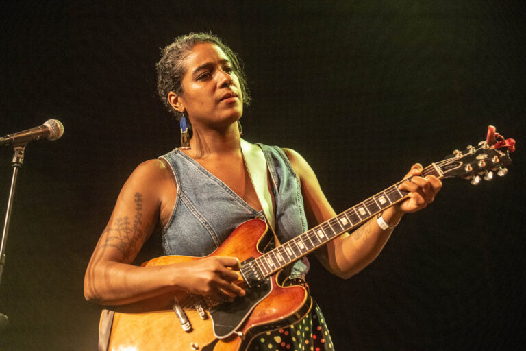 Een vrouw speelt gitaar op het podium, met een scherp licht op haar terwijl ze geconcentreerd naar het publiek kijkt.
