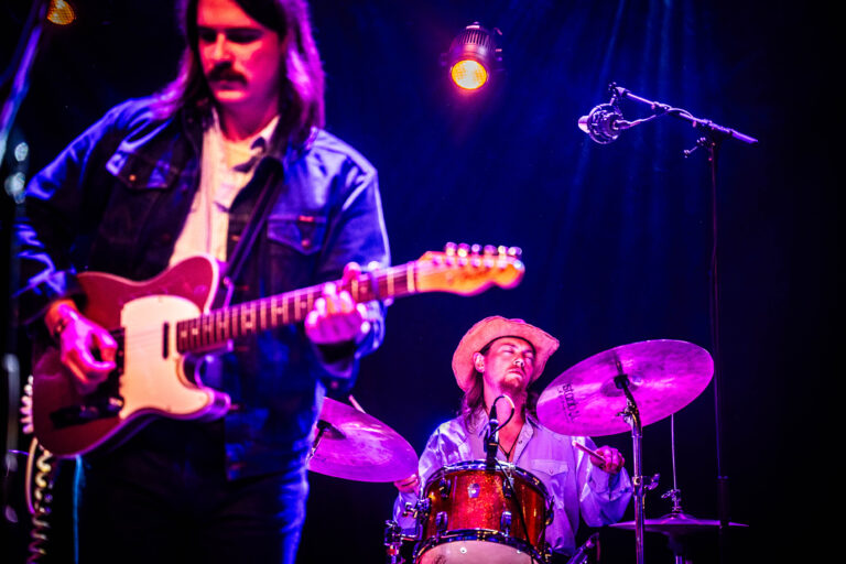 Twee muzikanten spelen op het podium: een gitarist met een elektrische gitaar en een drummer achter een set.