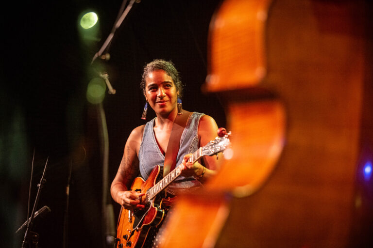 Een muzikante speelt gitaar op het podium, met een contrabas zichtbaar op de voorgrond.