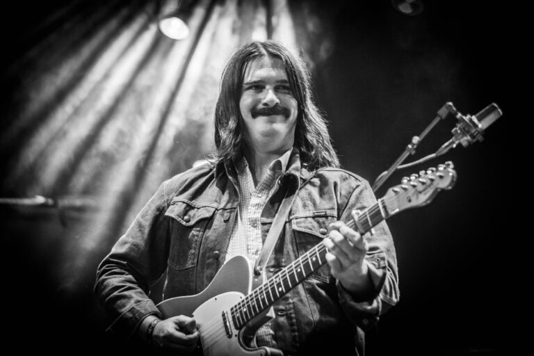 Een man met een snor speelt gitaar op het podium, omringd door lichtstralen in een zwart-wit foto.