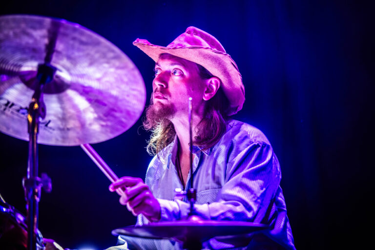Een drummer in een hoed speelt op een podium met paarse verlichting.