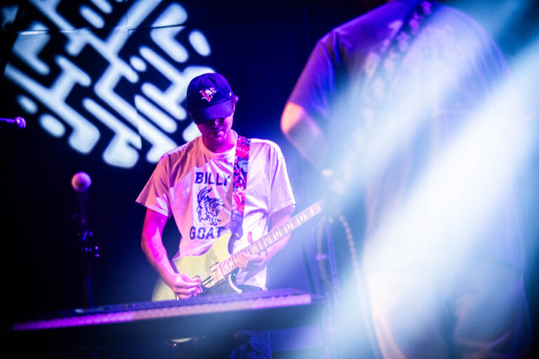 Een muzikant speelt gitaar op het podium, omringd door kleurrijke lichten.