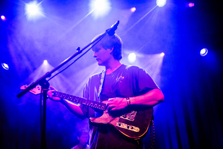 Een muzikant speelt gitaar op een podium, verlicht door paarse en blauwe lichten.