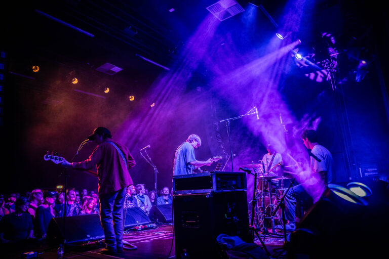 Een band speelt live op het podium, omgeven door kleurrijke lichten en een enthousiaste menigte.