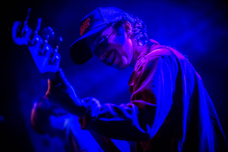 Een muzikant speelt gitaar op het podium, verlicht door paarse en blauwe lichten.