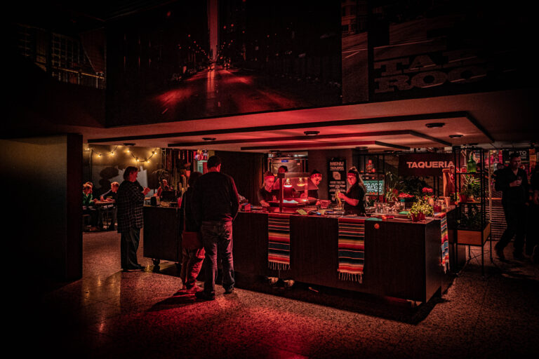 Een dimverlichte bar met een rode gloed, waar gasten aan het bestellen zijn en medewerkers aan het werk zijn.