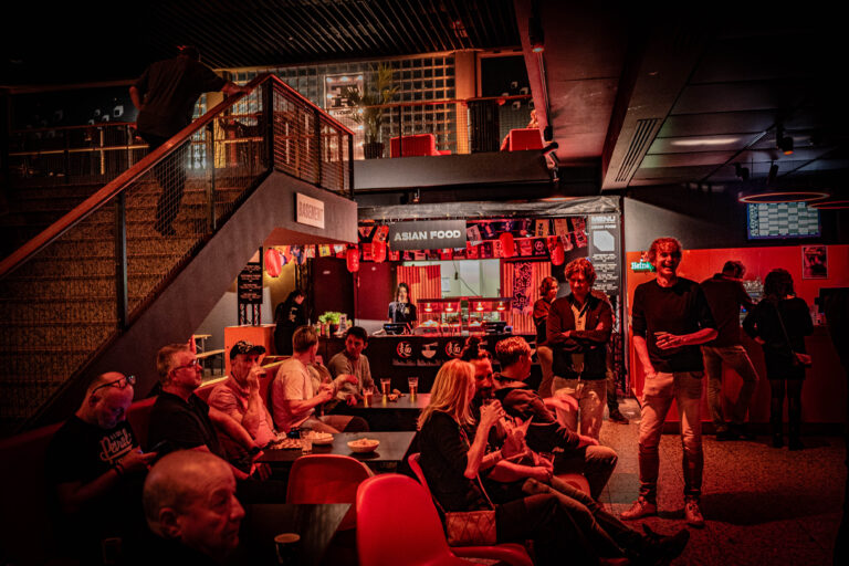 Een drukke bar met sfeerverlichting, waar mensen aan tafels zitten en een bar achter hen zichtbaar is.